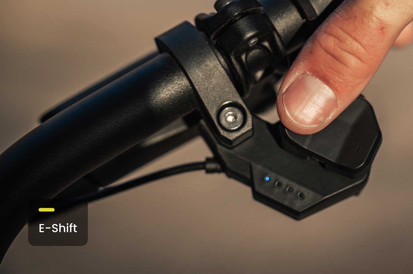 Aventon Aventure M ebike showing a Close-up of a hand operating an e-bike handlebar control with 'E-Shift' indicator. Shimano Cues electronic shifting available.