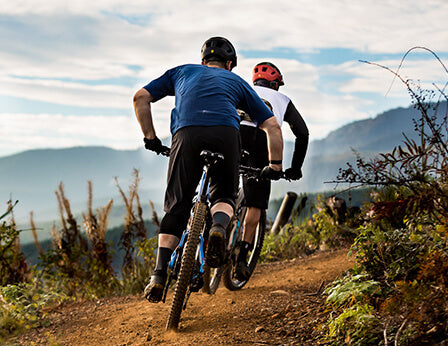 Two adults riding mountain bikes on a red dirt trail with beautiful summer mountain scenery